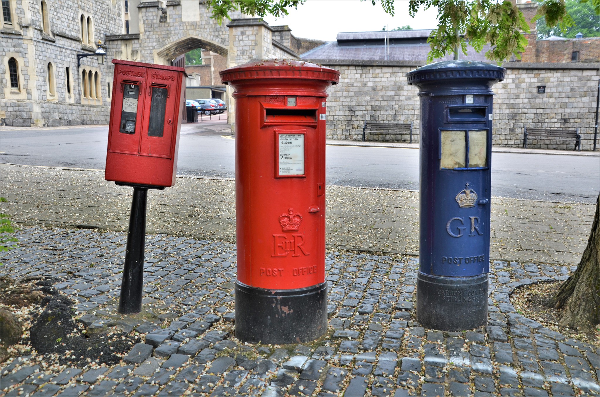 UK post boxes in a row: red stamp dispenser, red pillar box with ER cypher, and blue airmail pillar box with GR cypher on a cobbled pavement